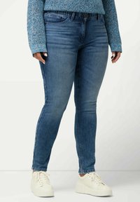 Jeans de mezclilla azul con un fit ajustado y rayas verticales. Presentan un cierre de botón en la parte frontal, bolsillos laterales y una textura suave. Llevados con zapatillas blancas.