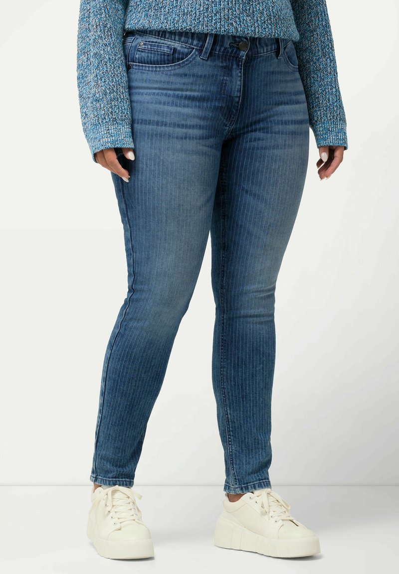 Jeans de mezclilla azul con un fit ajustado y rayas verticales. Presentan un cierre de botón en la parte frontal, bolsillos laterales y una textura suave. Llevados con zapatillas blancas.