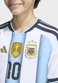 Osoba ubrana w narodową koszulkę piłkarską Argentyny w niebiesko-białe paski, z odznaką Mistrzów Świata FIFA 2022, numerem 10 oraz widoczną emblematyką AFA.