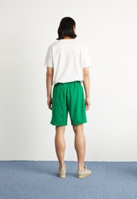 Gröna shorts med elastiskt midjeband, kombinerat med en enkel vit t-shirt och gråa slip-on skor, sedd bakifrån.