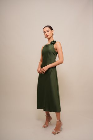 Frau in einem ärmellosen olivgrünen Midi-Kleid mit einem Schulterblüten-Detail und silbernen, schnürbaren High Heels steht vor einem beigen Hintergrund.