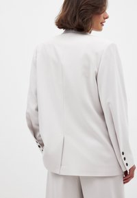 Gaiši pelēks bleizeris ar klasisku griezumu, kas aprīkots ar atvērumu aizmugurē un melnu pogu akcentiem uz piedurknēm. Gluda tekstūra.