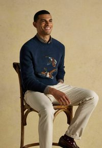 Naeratav mees istub puidust toolil, seljas tumesinine sviiter lennukite motiiviga, ruuduline särk, beezid püksid ja pruunid kingad, millel on mustrilised sokid.