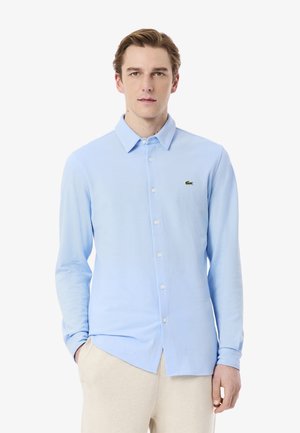 Camicia azzurra chiara con bottoni, realizzata in un tessuto morbido, con colletto classico e maniche lunghe, caratterizzata da un piccolo logo verde sul petto sinistro.