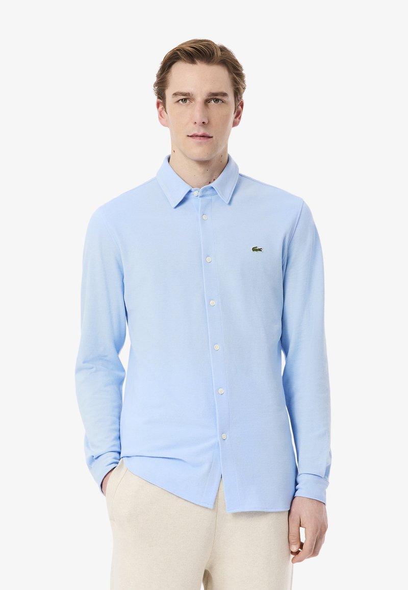 Camicia azzurra chiara con bottoni, realizzata in un tessuto morbido, con colletto classico e maniche lunghe, caratterizzata da un piccolo logo verde sul petto sinistro.