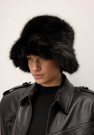 Melna kažokādas spaiņcepure ar platu, mīkstu malu un teksturētu apdari, valkāta ar melnu ādas jaku, kurai ir rāvējslēdzis un plecu detaļas.