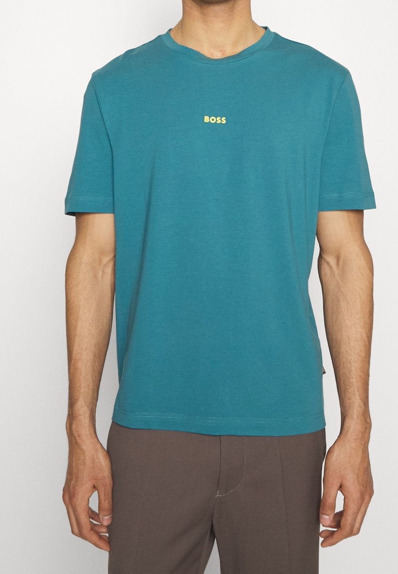 Homme portant un T-shirt à manches courtes teal avec un petit logo jaune "BOSS" sur la poitrine et un pantalon marron, sur un fond blanc.