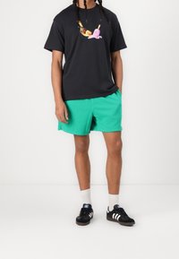 Camiseta negra con un gráfico colorido que presenta un pájaro y un helado, combinada con pantalones cortos verdes. Zapatillas negras con detalles blancos.