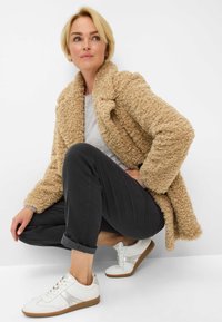 Beige, gestructureerde, oversized jas met een sjawl-kraag, grijze shirt, zwarte opgerolde broeken en witte sneakers met tan kleurige zolen.