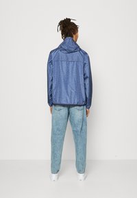 Blå mønstret jakke med høj krave, der har elastiske manchetter og en løs pasform. Lyseblå denimbukser fuldender looket.