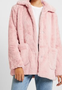 Rosa Kunstpelzmantel mit Kapuze, weicher Textur, zwei Fronttaschen und Druckknopfverschlüssen. Getragen über einem weißen Oberteil und blauer Jeans.