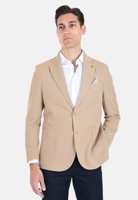 Blazer beige con texture, con rever a punta, due tasche frontali e un bottone dorato. Indossato sopra una camicia bianca con un fazzoletto da taschino a fantasia.