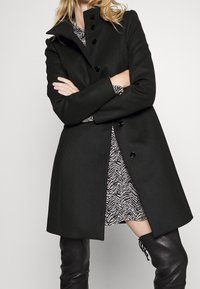 Cappotto lungo in misto lana nero con colletto alto, chiusura con bottoni e maniche lunghe. Indossato sopra un vestito a fantasia fino al ginocchio.