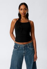 Schwarzes, enges Tanktop mit dünnen Trägern, kombiniert mit lockeren blauen Denim-Jeans, die eine ausgebleichte Textur und sichtbare Nähdetails aufweisen.
