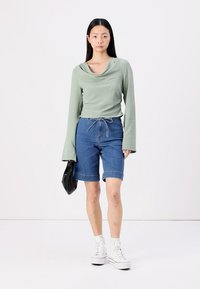 Top verde de manga larga con escote de cowl, combinado con pantalones cortos de mezclilla tipo Bermuda. La modelo sostiene un bolso negro y lleva zapatillas altas blancas.