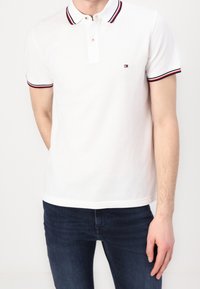 Vit polo med en tvåknaps placket, röd och marinblå randig krage och manschetter, korta ärmar och en liten logotyp på vänster bröst.