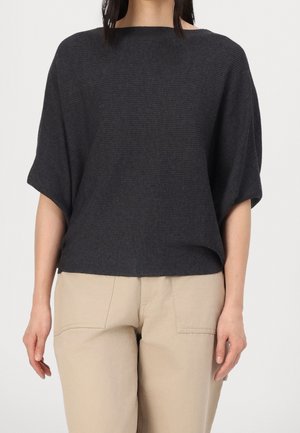 Woman wearing a loose dark gray ribbed short-sleeve top and beige pants, standing against a plain light background.