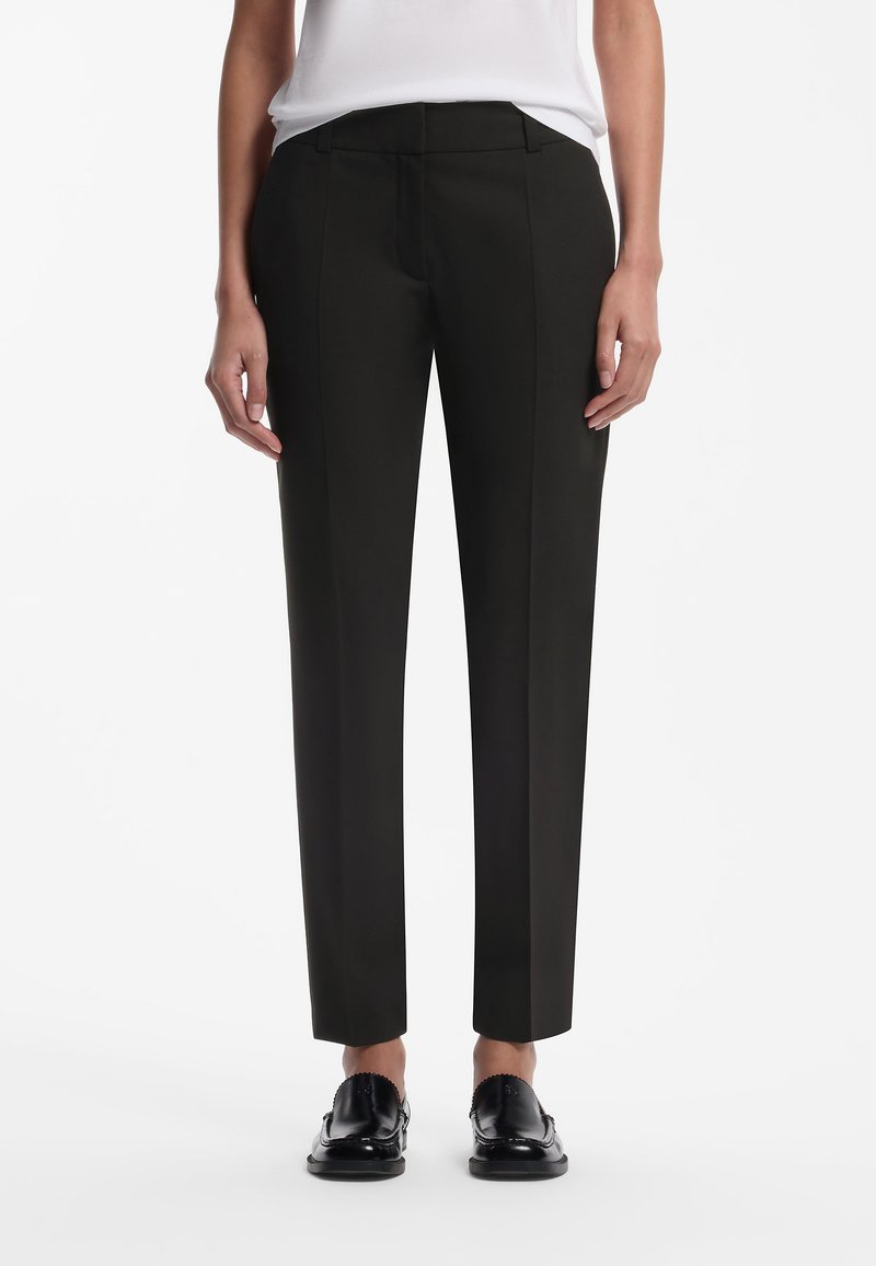 Femme portant un pantalon droit noir, un haut blanc et des mocassins noirs, debout sur un fond blanc uni.