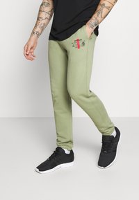 Gröna träningbyxor med en logotyptryck i svart och röd på vänster höft, med elastiska mudd och sidofickor. Kombineras med svarta träningsskor.