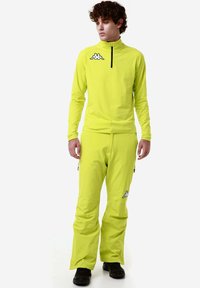 Top giallo brillante a maniche lunghe con colletto alto e zip; pantaloni abbinati con zip e dettagli del logo. Tessuto liscio e leggero.