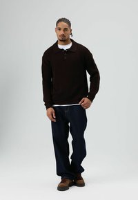 Jonge man met ingevlochten haar die een donkerbruine gebreide trui, wit ondershirt, losse donkere spijkerbroek en bruine laarzen draagt, staand tegen een effen achtergrond.