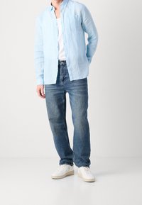 Camicia button-up in lino azzurro chiaro, top bianco sotto, indossata con jeans blu scoloriti e sneakers bianche. Design semplice, vestibilità rilassata.