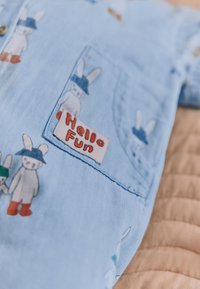 Svetlomodrá látka s vreckom so vzorom kreslených zajacov s klobúkmi a čižmami, jeden drží tabuľu s nápisom "Hello Fun."