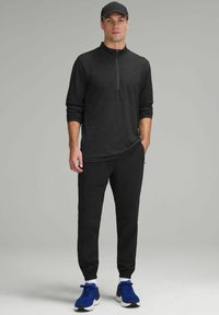 Schwarzes Langarmoberteil mit Reißverschluss und strukturiertem Muster, kombiniert mit schwarzen schmalen Hosen und blauen Sportschuhen.