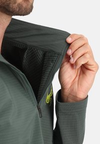 Veste verte avec un intérieur texturé, un col zippé et des manches. Présente un logo d'araignée jaune sur le côté. Tissu extérieur lisse.