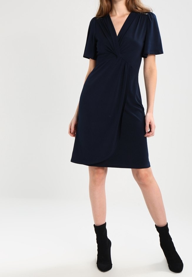 Robe bleu marine en tissu lisse, caractérisée par un col en V, des manches courtes et un détail froncé à l'avant. Portée avec des bottines noires.