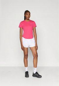 Różowy sportowy t-shirt z krótkim rękawem i okrągłym dekoltem, zestawiony z białymi szortami. Czarne buty sportowe i białe skarpetki z logo dopełniają look.