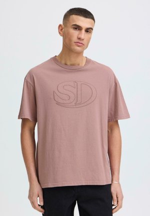 Joven vestido con una camiseta holgada color malva con el logo en relieve "SD" y pantalones negros, de pie frente a un fondo claro y liso.