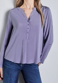 Maglietta a maniche lunghe e scollatura a V in tessuto lilla chiaro con bottoniera. Struttura liscia e leggero arricciamento sulle spalle. Abbinata a jeans blu scuro.