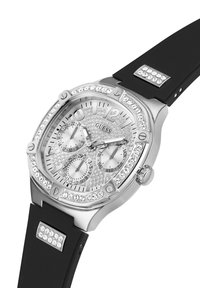 Strieborný hodinký s okrúhlym čelom, bezkompromisne osadeným krištáľmi, tromi subciferníkmi a čiernym gumovým remienkom. Obsahuje prvky z nerezovej ocele.