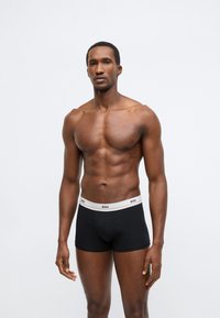 Boxeri negri cu talie albă și aurie pe care este imprimat logo-ul "BOSS". Materialul pare neted și ajustat. Modelul stă într-o poziție neutră.