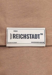 Tessuto marrone con motivo a maglia strutturata, con un'etichetta bianca che include "REICHSTADT", istruzioni per la cura e dettagli del sito web.