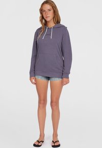Paarse hoodie met trekkoorden, een voorkauwzak en ribbelmanchetten; gedragen over denimshorts en zwarte teenslippers. Gladde stoftextuur.