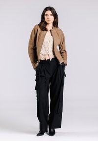Giacca bomber in suede marrone, top in pizzo crema, pantaloni cargo neri con tasche e tacchi a punta neri. Colori solidi e materiali testurizzati.