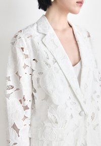 White lace blazer featuring floral patterns, a notched lapel, and a single button closure. The fabric is textured and semi-sheer.