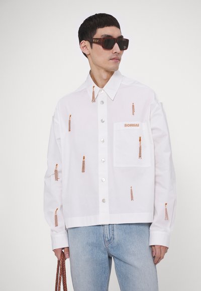 Hvid langærmet skjorte med orange broderede mønstre, knaplukning og en brystlomme; parret med lyseblå jeans og solbriller.