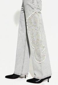 Lysegrå brede jeans med slidser på låret og et kontrasterende broderet design. Kombineret med spidse sorte hæle.