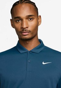 Nike polo tričko v tmavě modré barvě, s klasickým límcem, dvoubarevným zapínáním na knoflíky a bílým logem na levé části hrudi. Hladká, texturovaná tkanina.