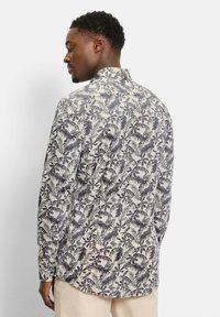 Mann trägt ein beige langärmliges Hemd mit dunkelgrünem botanischem Blattmuster, steht seitlich und blickt leicht nach unten.