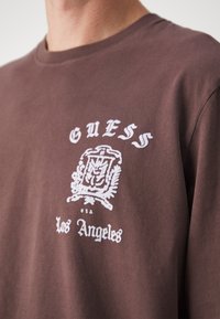 Maglietta di cotone marrone con maniche corte. Presenta un logo stampato bianco e la scritta "GUESS Los Angeles," con un emblema decorativo sopra il testo.