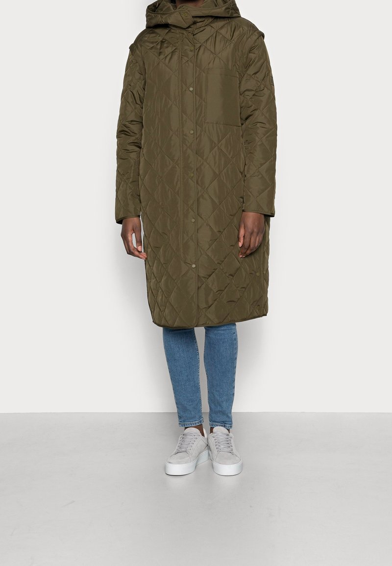 Cappotto trapuntato verde oliva con cappuccio, lunghezza media, con tasca frontale e bottoni automatici. Indossato sopra jeans blu e sneakers bianche.