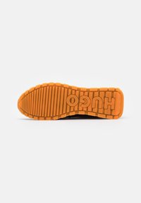 Suola di sneaker nera con fondo in gomma arancione testurizzata, dotata di scanalature rialzate e con il nome del marchio "HUGO" ben visibile al centro.