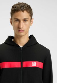 Felpa con zip nera, con una striscia orizzontale rossa su cui è stampato "HUGO" in testo bianco. Ha un cappuccio e un tessuto liscio e strutturato.