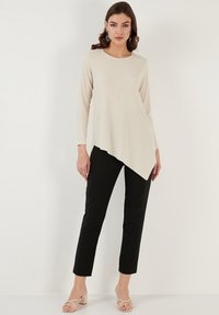 Top beige asimmetrico a maniche lunghe con superficie testurizzata, abbinato a pantaloni neri aderenti. Il modello indossa sandali a strappi di colore chiaro.