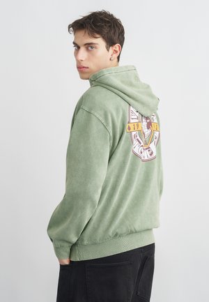 Young man with short brown hair wearing a faded green hoodie with a graphic design on the back and black pants, turned slightly to the side.