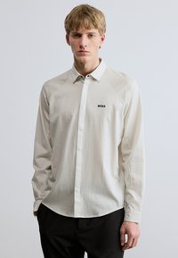 Ilgas rankovės marškiniai šviesiai kreminės spalvos su subtiliomis vertikaliomis juostelėmis ir juodu „BOSS“ logotipu kairėje krūtinės pusėje. Lygus audinys; prigludusi iškirptė.